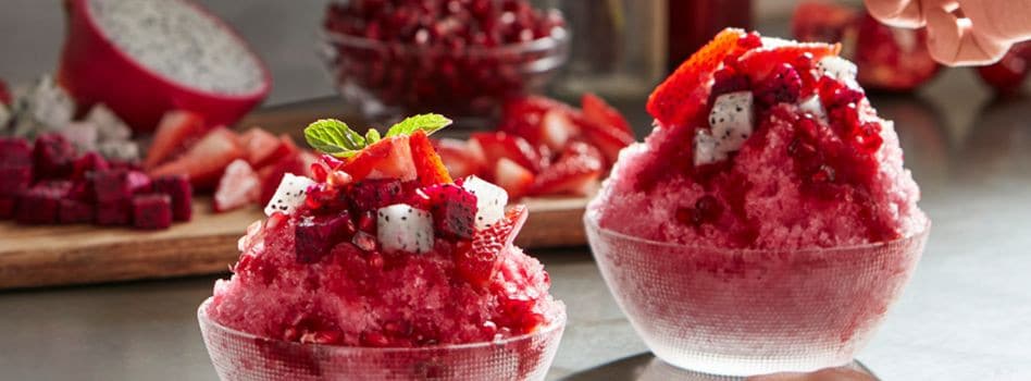 Is Hawaiian Shave Ice Different from Snow Cones A hand gently places mint leaves onto the surface of delicious shaved ice treats, which is coated in red syrup and accompanied by slices of fresh fruit."