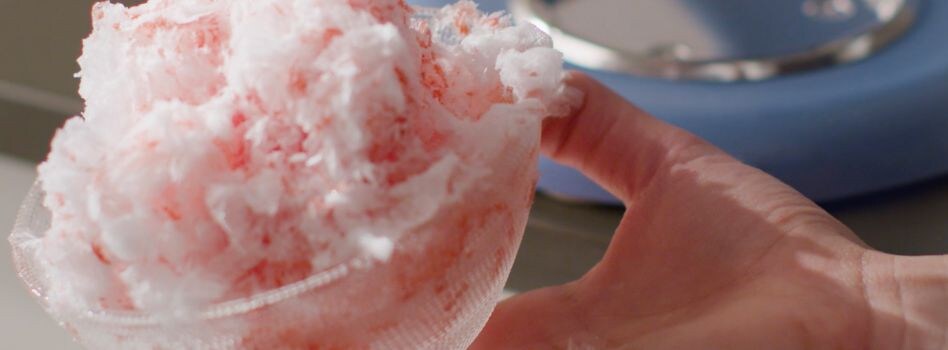 Is Shaved Ice As Crunchy As Snow Cones Une main tient un récipient en verre rempli de glace pilée, arrosée de sirop rouge pour donner de la saveur.