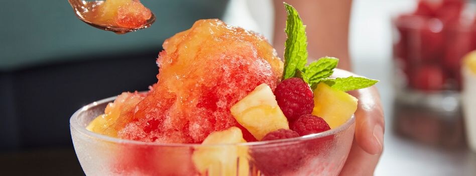 How to Make Shaved Ice at Home Une main tient un bol en verre transparent contenant de la glace pilée de couleur rouge et orange, garnie de fruits frais et de feuilles de menthe.