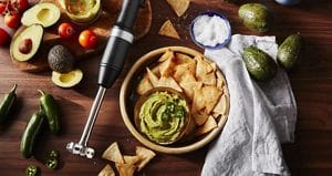 On a wooden surface are cut avocadoes some with the pits still in, whole avocadoes, jalapeno peppers, a KitchenAid immersion hand blender, pita chips, a bowl of salt, and a bowl of pita chips and guacamoles.