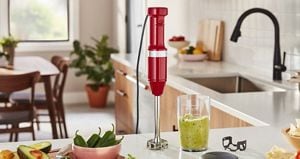 A red KitchenAid immersion hand blender stands upright on a counter. Next to it is a glass filled with guacamole, a bowl of jalapenos and some avocadoes sliced in half.
