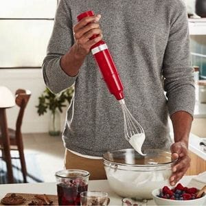 A person in a grey shirt and beige pants makes whipped cream with an immersion hand blender and whisk attachment. In front of them is a bowl filled with strawberries and blueberries and a cutting board with two glasses on top. In the background is a chair.