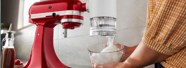 Someone catching ice in a bowl from the shave ice attachment of a red KitchenAid Stand Mixer.