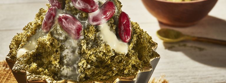 Du bingsu recouvert de poudre de matcha, de haricots rouges et de lait concentré. À côté se trouve un bol de matcha. 