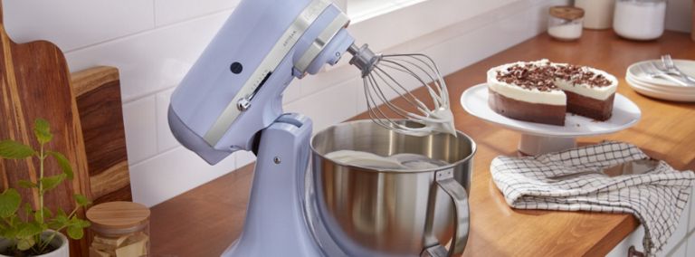 A KitchenAid Stand Mixer with the head tilted. The bowl is filled with a white mixture. Next to it is a cake on a cake dish and a dish cloth.