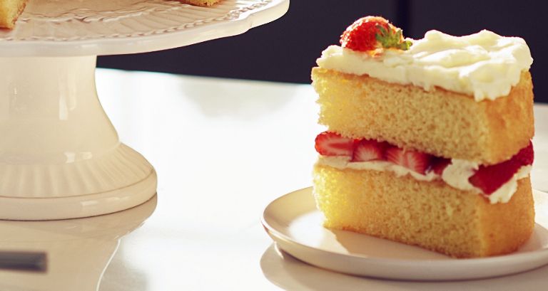 A piece of cake with white frosting and strawberries on a plate on a counter. A cake dish is next to it.