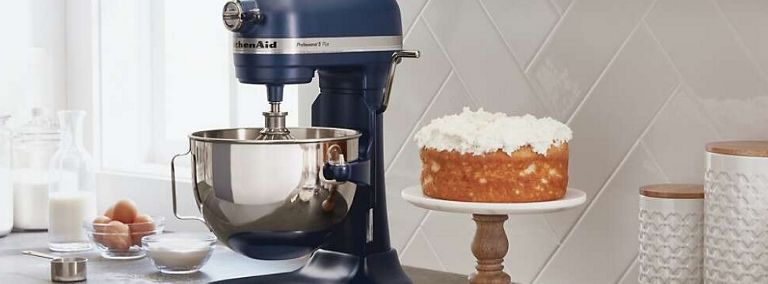 A dark blue KitchenAid Stand Mixer is on a counter. Also on the counter is a cake with white frosting on a cake dish, a bowl of eggs, a bowl with a white ingredient, two white containers and a container of milk.