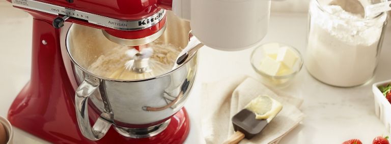 Un batteur sur socle KitchenAid rouge, équipé de l'accessoire balance et tamis, en train de battre et de mélanger des ingrédients. À côté de lui se trouvent un bol et un linge à vaisselle.