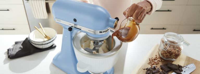 Quelqu'un verse un liquide dans le bol de congélation d'un batteur sur socle KitchenAid bleu. Sur le comptoir se trouvent également une planche à découper avec un bocal d'ingrédients, un couteau et du chocolat sur le dessus ainsi qu'un bol contenant deux cuillères sur un linge à vaisselle.