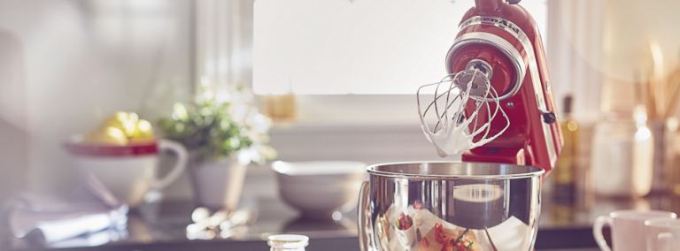 A red KitchenAid Stand Mixer with the head tilted up. Behind it are a bowl, a plant and a window.