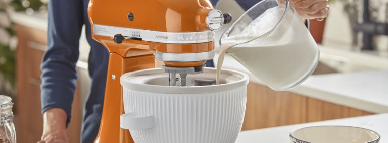 Someone in a blue shirt pours milk or cream into the bowl of a KitchenAid Stand Mixer. Next to the appliance are two stacked bowls and a dish cloth.