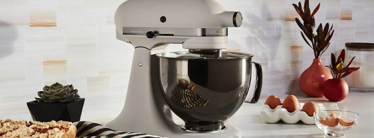 On a counter is a white KitchenAid Stand Mixer. Also on the counter are plants, eggs in a carton, a loaf of bread with a few slices in front, a dish cloth and a small bowl with eggshells.