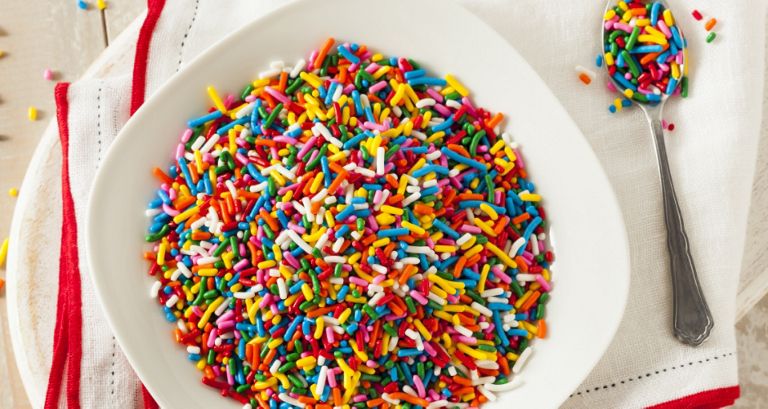 A bowl of rainbow sprinkles on a red and white dish cloth. Next to it is a spoon with sprinkles. 