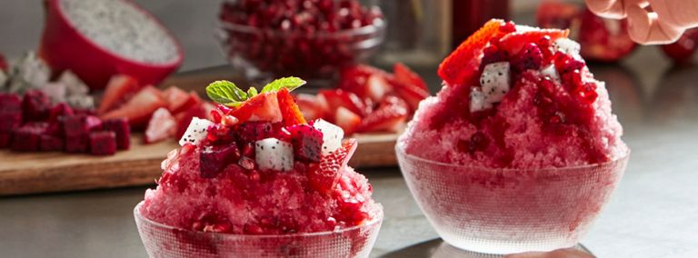 Une personne garnit un cône de glace rouge avec du basilic. Le cône est garni de grenades, de fraises et de fruit du dragon. Un autre cône similaire se trouve à côté. En arrière-plan, on voit des fraises coupées sur une planche à découper, la moitié d'un fruit du dragon et un bol de grenades.