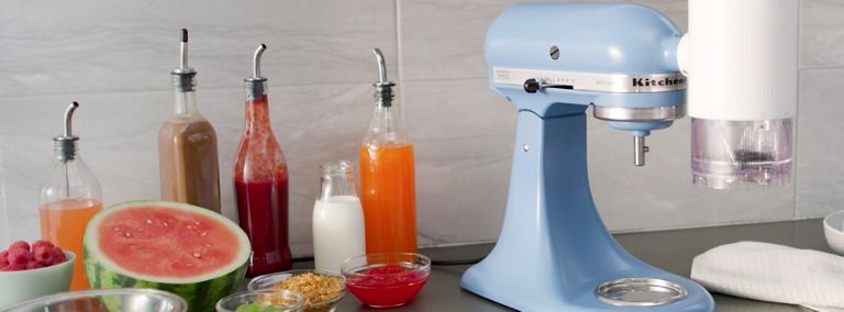 Un batteur sur socle KitchenAid bleu clair avec l' accessoire pour glace pilée se trouve sur un comptoir. Sur le comptoir se trouvent également des bols de garnitures, un demi-melon d'eau, un récipient de lait et plusieurs récipients sirops brun, rouge et orange. 