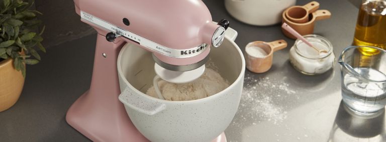 On a counter is a pink KitchenAid Stand Mixer with dough in its bowl. Also on the counter are measuring cups, including one with flour and one with water, a container of oil, a container of flour and a plant. Some flour dusts the counter.