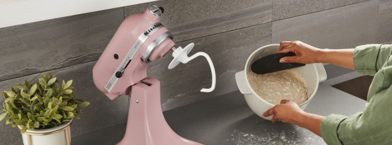  Someone scrapes dough out of a stand mixer bowl. Next to them is a pink KitchenAid Stand Mixer and a plant. 