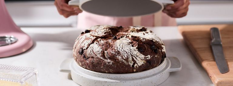  On a counter, someone unveils a rye bread. Next to the bread is a cutting board with a knife on top.