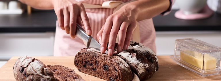 A person in a purple checkered shirt and apron slices rye bread on a counter. On the same cutting board as the bread is a stick of butter in a clear container.