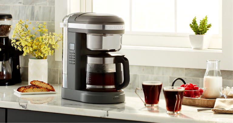 gray KitchenAid coffee maker surrounded by cups of coffee, cream, and other items.
