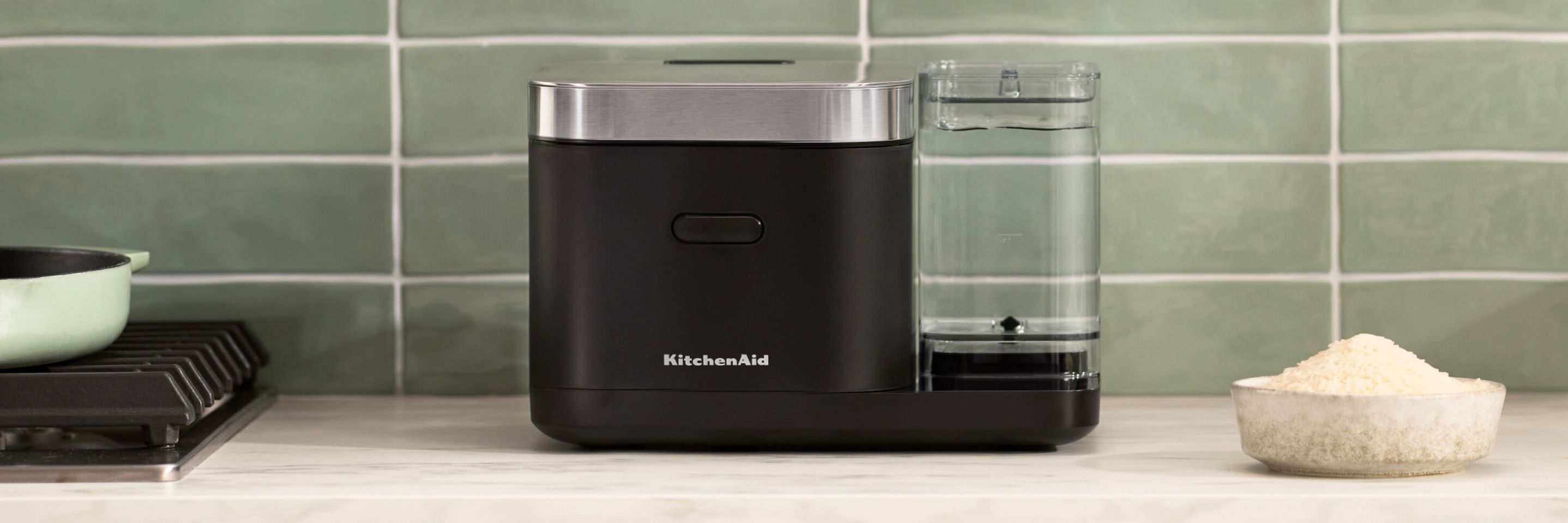 The KitchenAid® Grain & Rice Cooker sitting on a countertop with a green backsplash behind it.