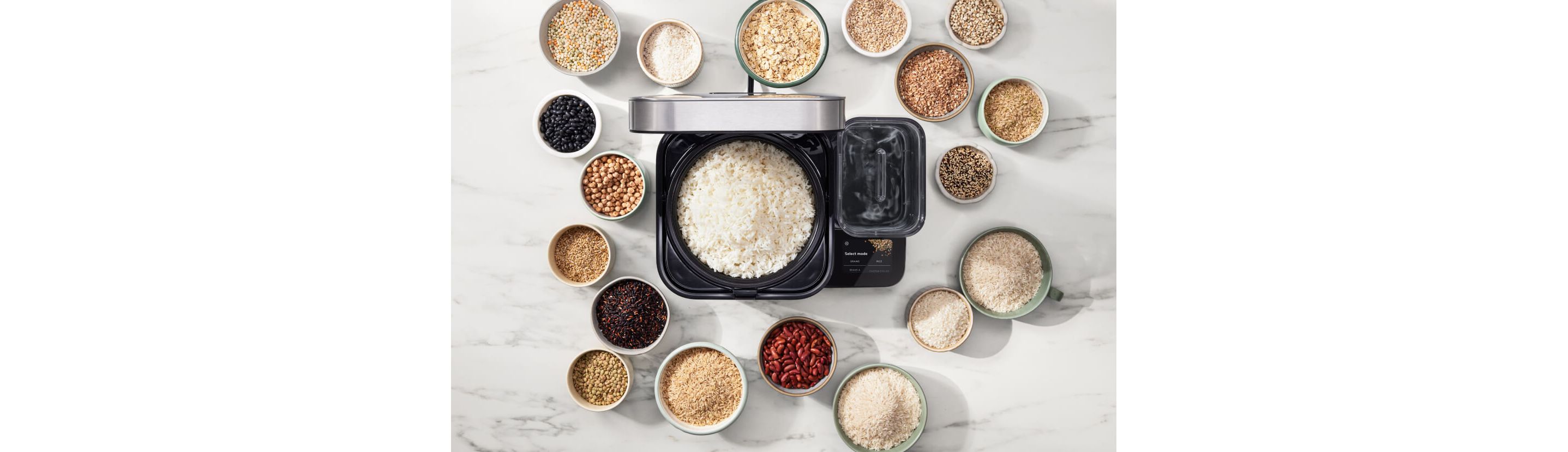 The open rice and grain cooker, with cooked white rice inside. The cooker is surrounded by bowls of various grains, rices and beans.